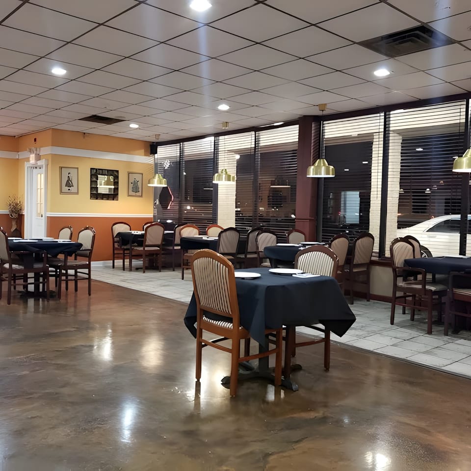 Empty restaurant dining room with tables set with dark tablecloths and chairs, illuminated by overhead lights.