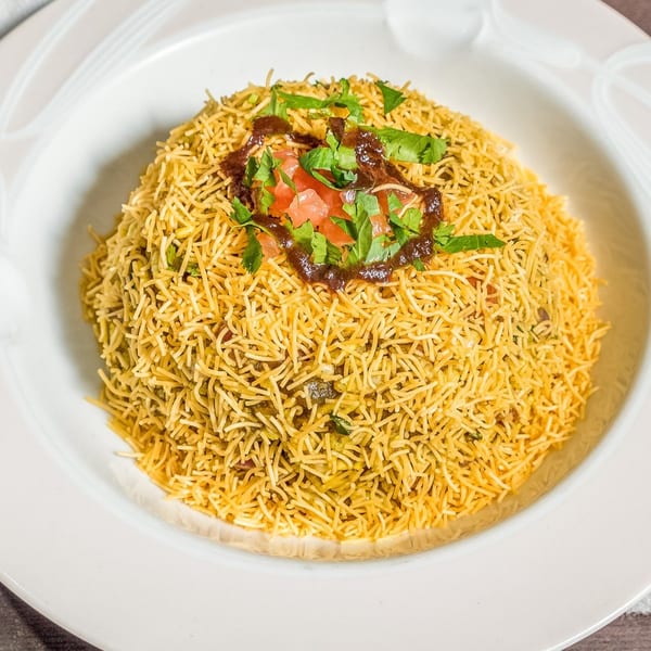 A white bowl filled with golden Indian sev, topped with chutney, diced tomatoes, and fresh cilantro.