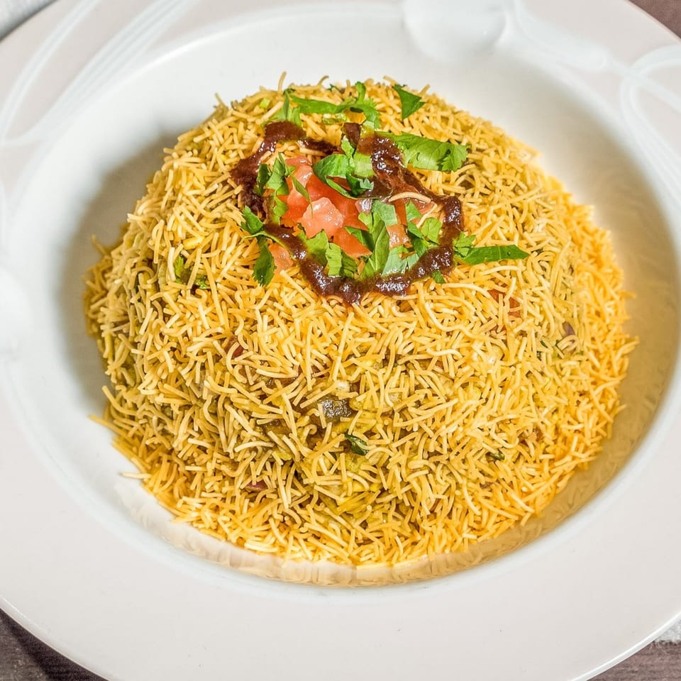 A white bowl filled with golden Indian sev, topped with chutney, diced tomatoes, and fresh cilantro.