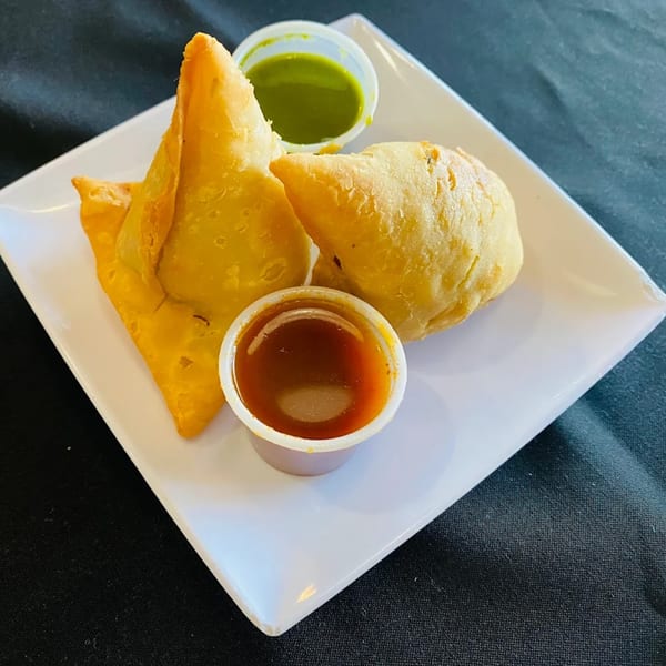 Two golden-brown Indian samosas with small cups of green and brown dipping sauces on a white plate.