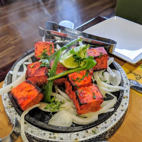 Steaming paneer tikka, garnished with onions, green peppers, and lime, served on a hot plate.